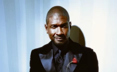 USHER in black suit with red lapel pin, standing against striped wall.